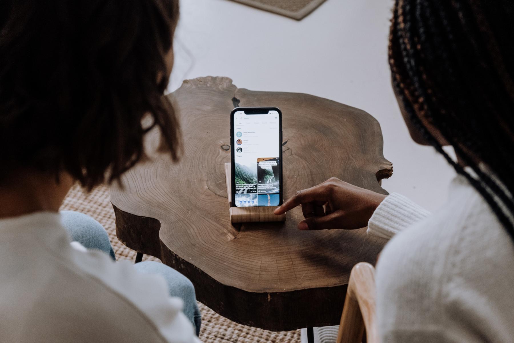 2 people watching a phone on a small wooden table