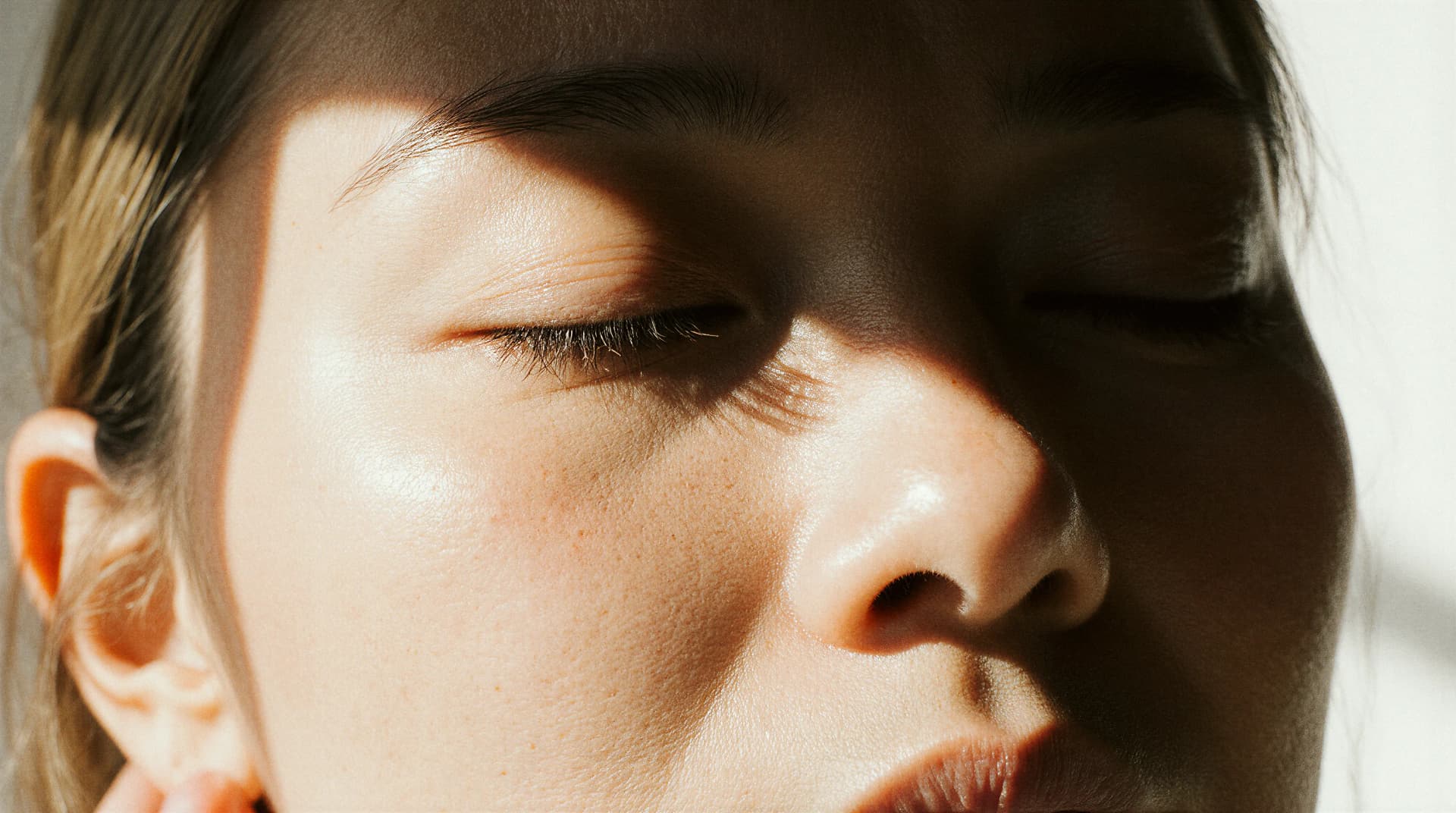 Close-up of a person's face with closed eyes, bathed in sunlight, casting soft shadows across their skin.