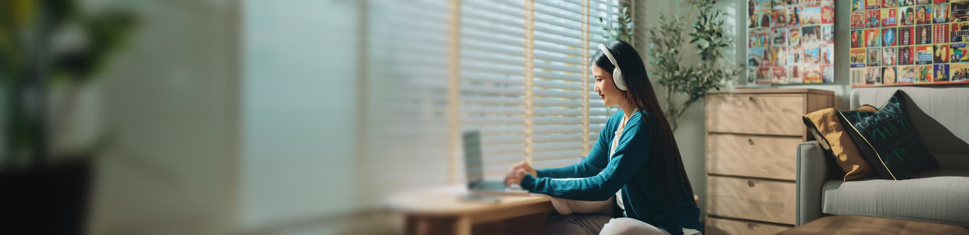 A woman sits by a window working on a laptop, wearing headphones. The room is cozy with plants, colorful artwork, and decorative pillows on a sofa.