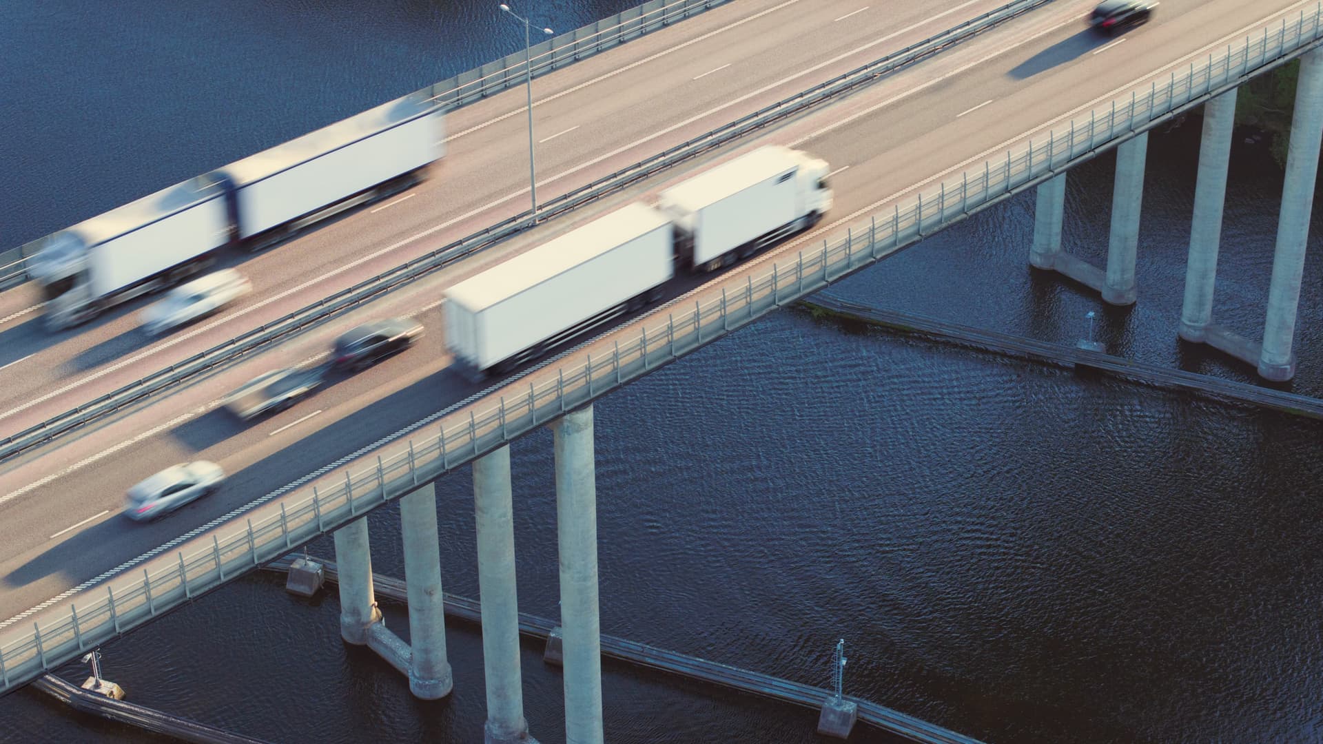 Cars and trucks drive over a bridge spanning a body of water, supported by tall concrete pillars, with blurred motion suggesting speed.