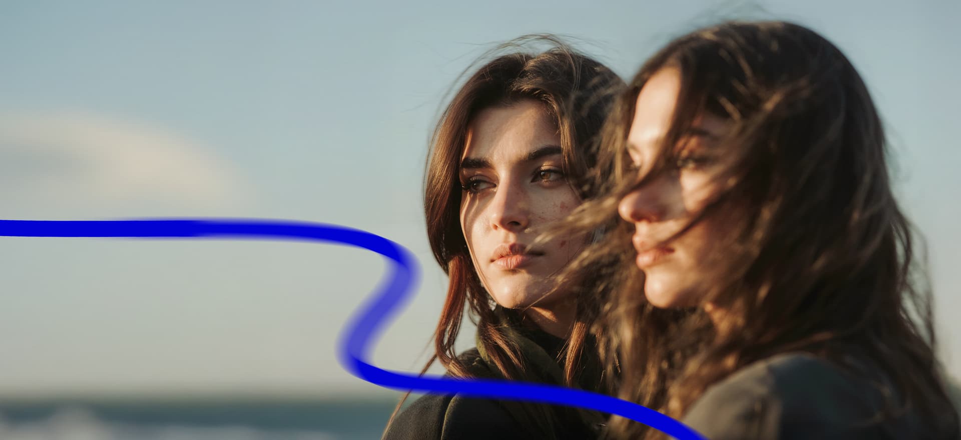 Two women with windblown hair standing outdoors, looking pensive. A blue wavy line appears across the image.