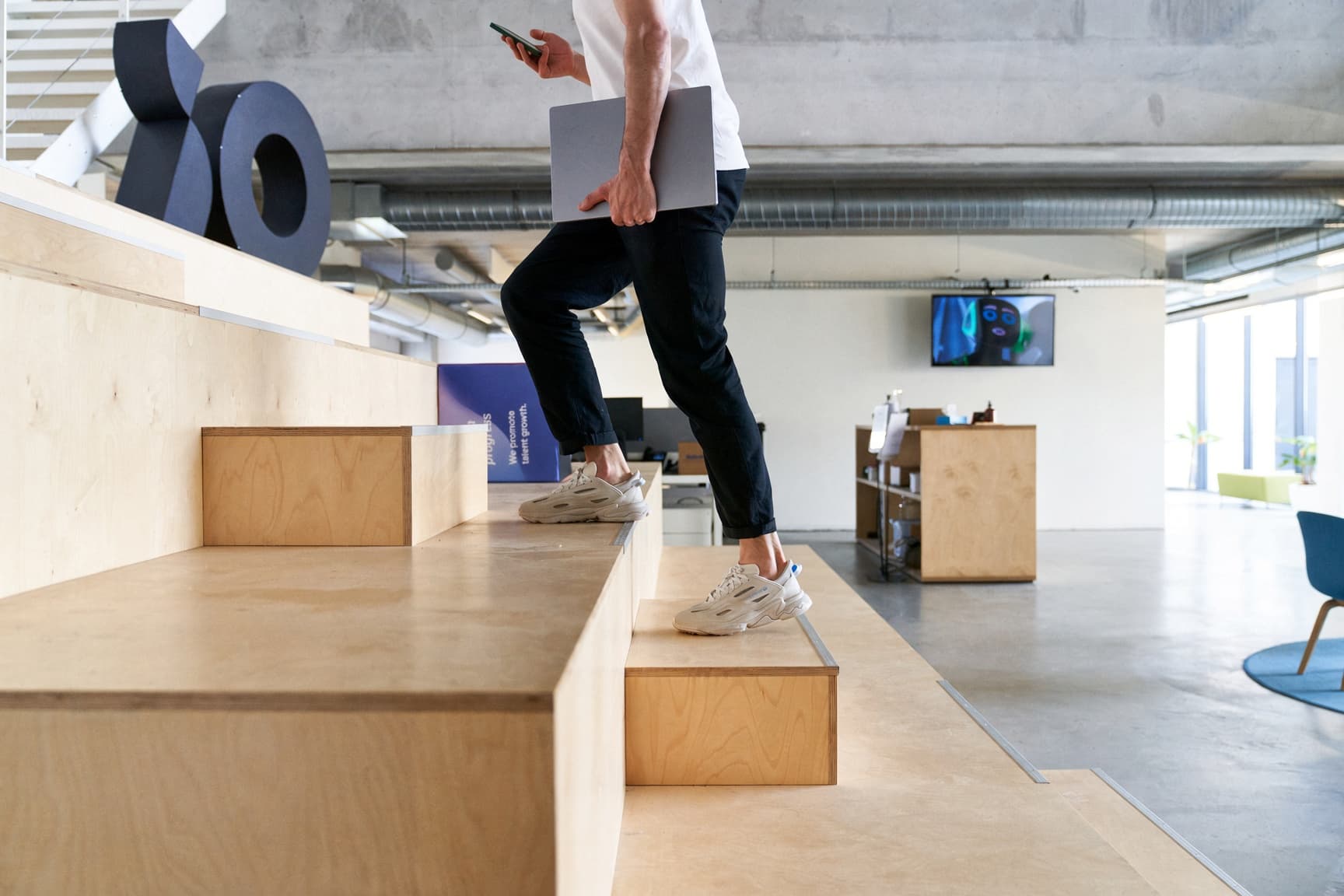 A person walking up wooden steps in a modern office, holding a laptop and phone, wearing casual clothing and sneakers.