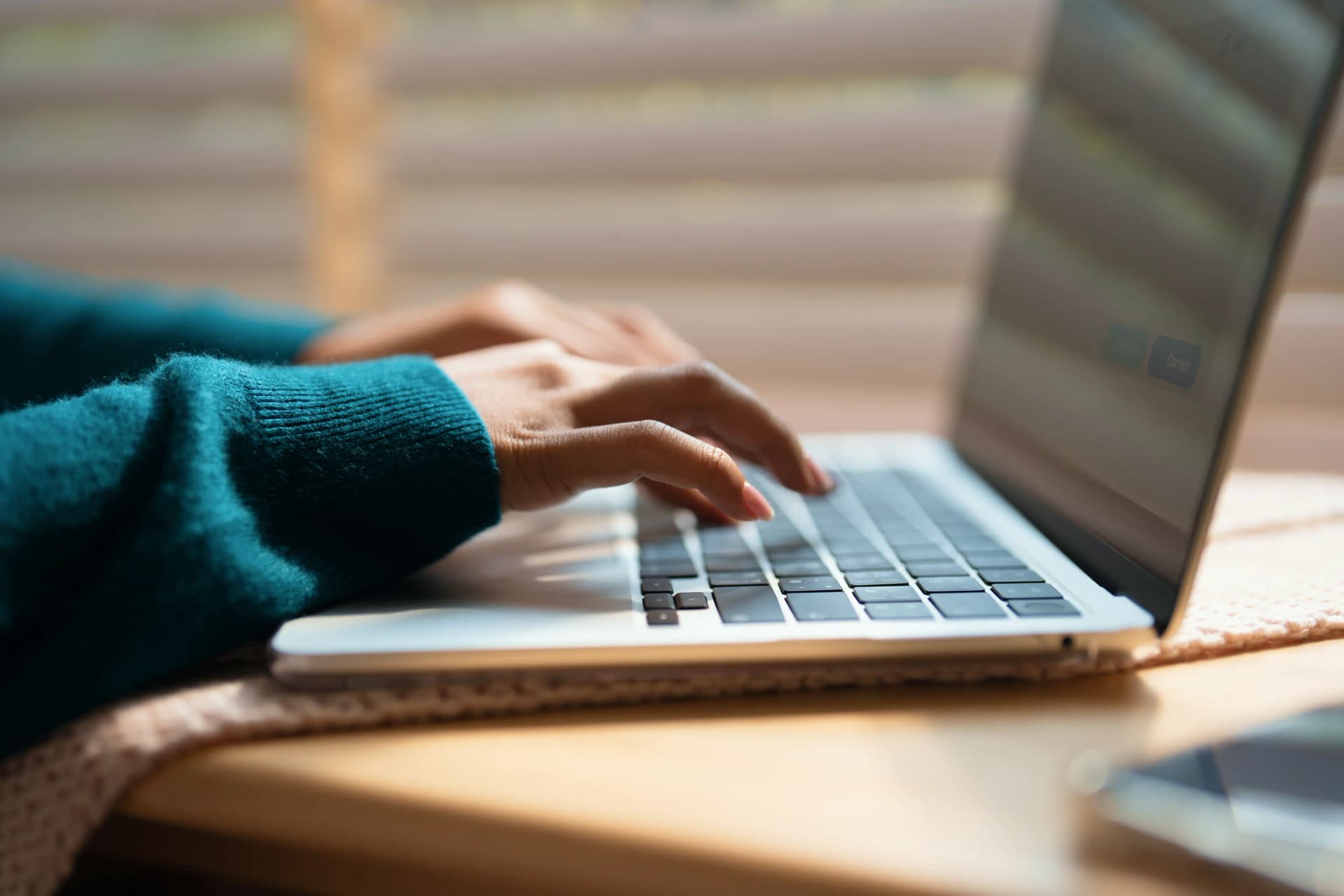 Person typing on a laptop, wearing a teal sweater, with a blurred background of window blinds and a table.