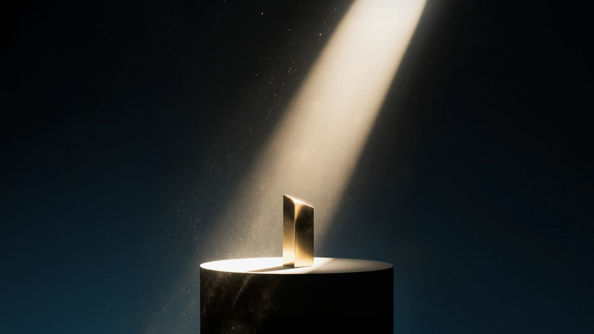 A gold, abstract sculpture on a round black pedestal is illuminated by a spotlight in a dark room, creating a dramatic effect.